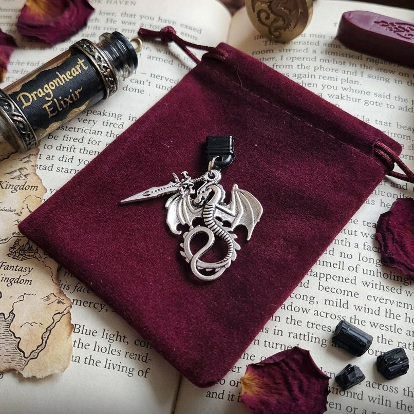 A silver dragon and sword pendant displayed on a deep burgundy velvet drawstring pouch, styled on an open book with dried rose petals, a bottle labeled Dragonheart Elixir, and fantasy-themed props. Text at the bottom reads 'The Dragon & Sword Collection - The Book Haven Shop'.